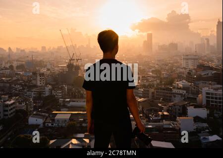 Giovane asiatico con macchina fotografica in cima all'edificio e vista dell'alba sulla città di Bangkok in Thailandia Foto Stock