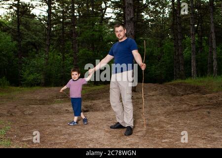 Papà e figlio camminano nella foresta di pini conifere la sera e giocano con bastoni. Festa del Padre, il ruolo del papà nella vita del bambino. Foto Stock