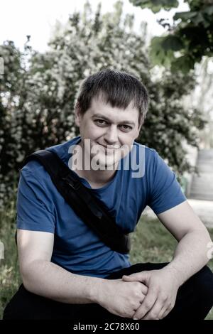 Un uomo, 25 e 30, che indossa una T-shirt blu con uno zaino si siede nel selvaggio vicino alberi. Foto Stock