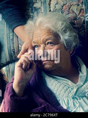 Anziani pene Sad Care Senior Lady 90 anni a casa con espressione sonnolente grave mantiene tattile confortante mano affidabile di caregiver in luce naturale interni Ritratto verticale retro stile Foto Stock