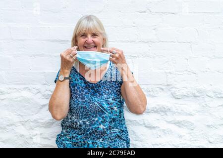 Donna sorridente, indossando una maschera Coronavirus, pandemia Covid-19, pronta per la fine del blocco, indossando quando fuori in pubblico. Sfondo bianco chiaro. Foto Stock
