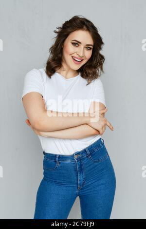 Ragazza positiva in jeans, maglietta bianca e paletti rossi che si posano vicino alla parete tenendo le mani incrociate sul petto Foto Stock