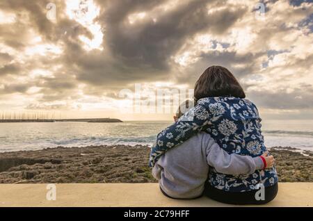 Mamma con figlio seduto e abbracciato l'un l'altro sul muro che guarda al mare Foto Stock