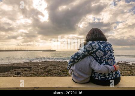 Mamma con figlio seduto e abbracciato l'un l'altro sul muro che guarda al mare Foto Stock