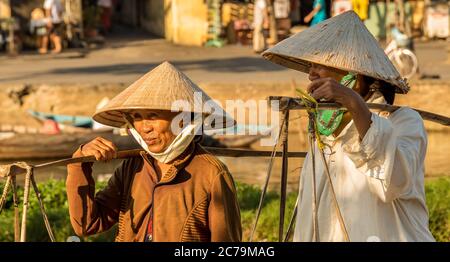 Due donne vietnamite con cappelli conici, che portano le spalle di un fiume nel pomeriggio, Hoi An, Vietnam - 10 gennaio 2015 Foto Stock