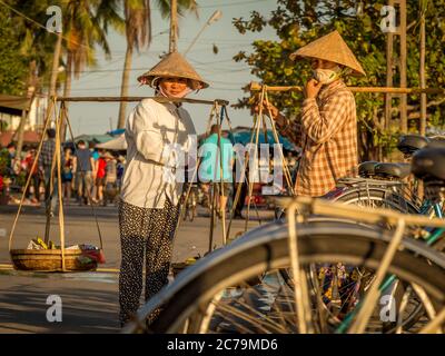 Due donne vietnamite con cappelli conici, che portano le spalle di un fiume nel pomeriggio, Hoi An, Vietnam - 10 gennaio 2015 Foto Stock