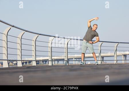 Uomo maturo atletico in sportswear lato curvante con braccio rialzato mentre fa riscaldare su terrapieno Foto Stock