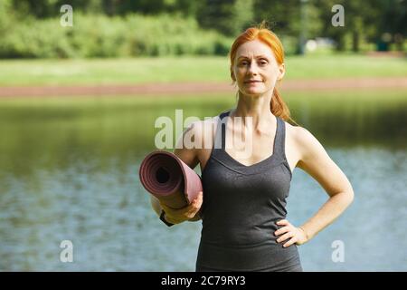 Ritratto di una donna matura e sicura che si trova contro il lago e che tiene un tappetino yoga Foto Stock