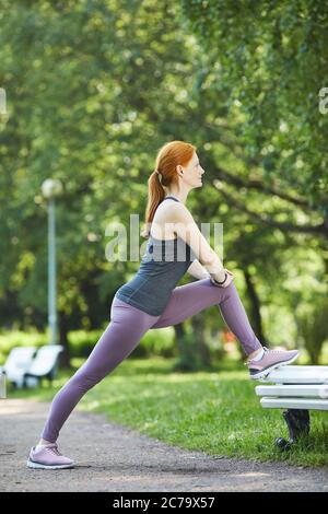 Vista laterale della donna sottile e matura con coda di pontile poggiata sul piede sulla panca mentre si fa esercizio stretching nel parco cittadino Foto Stock