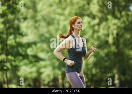 Donna rossa seria matura in auricolari wireless che indossano il tracker di fitness che corre nel parco verde Foto Stock