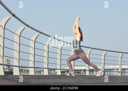 Donna di mezza età sportiva con occhi chiusi che eseguono la posizione guerriero con braccia sollevate sull'argine della città Foto Stock