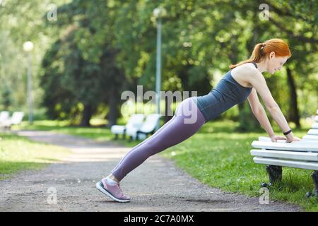 Vista laterale di una donna robusta e matura in testa rossa con leggings che fanno push-up dalla panchina nel parco cittadino Foto Stock