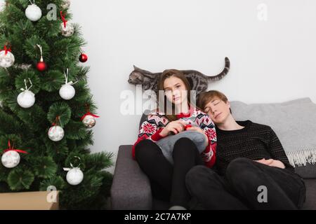 Ritratto di bella ragazza seduta su divano e sognante guardando in macchina fotografica mentre ragazzo si appoggia su di lei e dormire con gatto e albero di Natale su Foto Stock