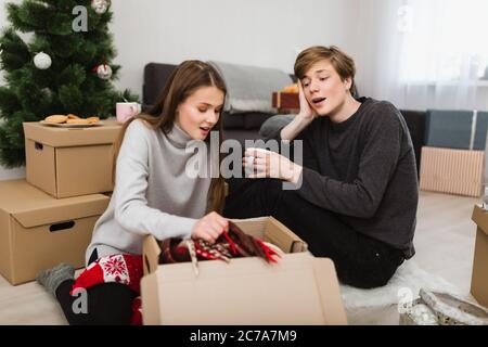 Ritratto di bella giovane coppia seduta sul pavimento a casa e stupefatto guardando in scatola con albero di Natale sullo sfondo Foto Stock