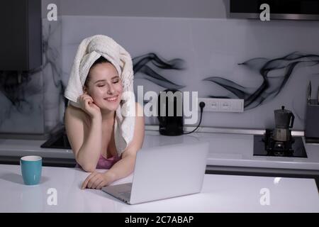 allegra bella bruna ragazza - in pigiama rosa e con un asciugamano bianco sulla testa - con una tazza blu tè-caffè, funziona su un computer portatile - lavoro remoto o studio - in una cucina grigia. Foto di alta qualità Foto Stock