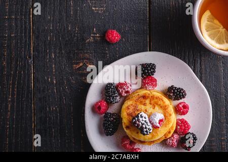 Frittelle di formaggio cottage, frittelle di cagliata dessert con lampone e frutti di bosco in piatto vicino alla tazza di tè caldo con fetta di limone Foto Stock