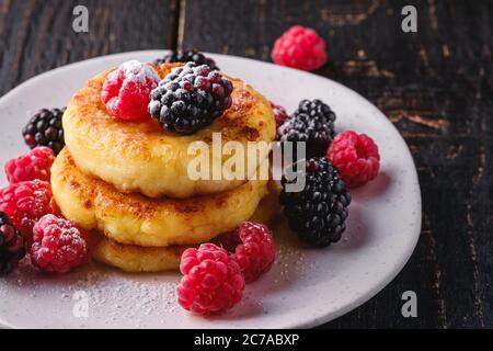 Frittelle di formaggio e zucchero in polvere, frittelle di cagliata dessert con lampone e frutti di bosco in piatto su sfondo nero scuro di legno Foto Stock