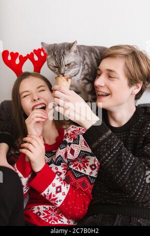 Ritratto di giovane coppia fresca seduta sul divano a casa con gelato in mani e felice di passare il tempo insieme mentre si gioca con gatto Foto Stock