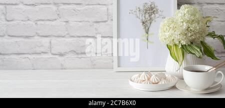 Tea in una tazza con un setaccio dorato. Bizet su un piatto. Cornice con fiori secchi. Sfondo mattone bianco invecchiato. Idrangea verde in un vaso nei paesi nordici Foto Stock