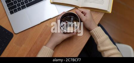 Vista dall'alto delle mani femminili che tengono una tazza di caffè ghiacciato su un tavolo di lavoro in legno con computer portatile e libro Foto Stock