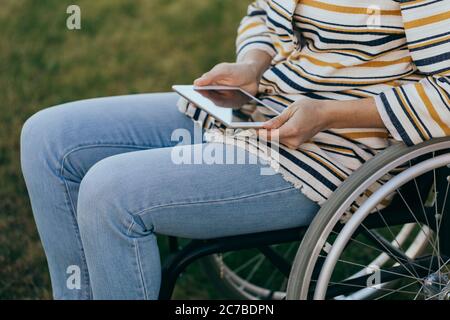 Donna vestita alla moda in sedia a rotelle con un moderno tablet in mano cammina per strada. Persona irriconoscibile. Foto Stock