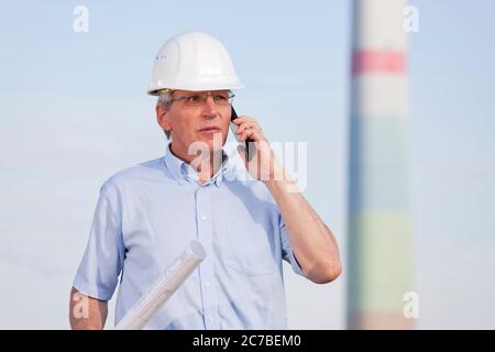 Ingegnere o architetto con un progetto in mano su un cantiere che parla sul telefono cellulare Foto Stock