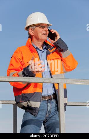 Caposquadra con cappello e abbigliamento protettivo che parla sul telefono cellulare su un sito di costruzione - Focus sull'uomo Foto Stock