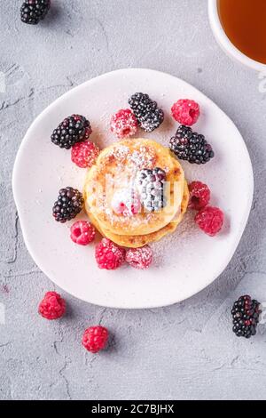 Frittelle al formaggio e zucchero in polvere, frittelle di cagliata dessert con lampone e frutti di bosco in piatto vicino alla tazza di tè caldo con fetta di limone Foto Stock