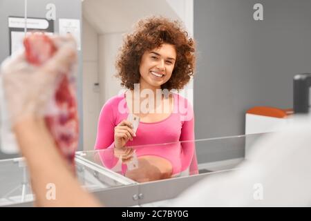 Fuoco selettivo del macellaio maschio incognito che tiene il pezzo di carne fresca rossa cruda e che mostra alla giovane donna. Vista frontale della signora con capelli ricci che tengono la carta di debito in negozio. Foto Stock
