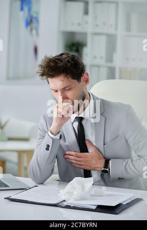 Uomo d'affari malato con barba seduta alla scrivania con documenti e tenendo la mano sul petto mentre si verificano sintomi di tosse, coronavirus o polmonite Foto Stock