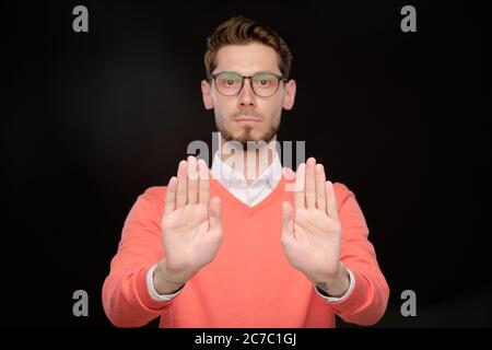 Serio stretto giovane uomo sopportato in occhiali gesturando le mani mentre chiede di smettere di muoversi, sfondo nero Foto Stock
