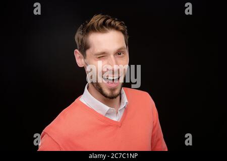 Ritratto di un giovane allegro e giocoso con barba che si inocchiava con la fotocamera su sfondo nero Foto Stock