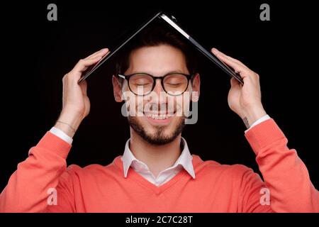 Giovane sorridente e spensierato con occhi chiusi che tengono il laptop sopra la testa mentre si nasconde al lavoro Foto Stock