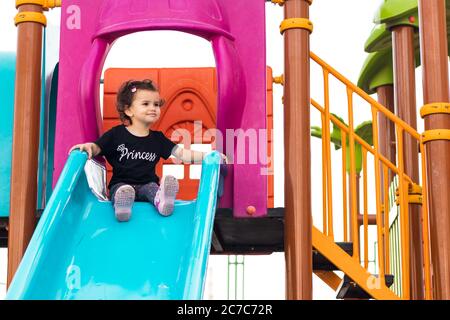una ragazza liitle carina seduta sullo scivolo blu in un parco giochi Foto Stock