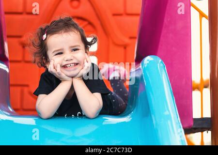 una ragazza liitle carina che si posa sullo scivolo blu in un parco giochi con le mani sotto il mento. Foto Stock