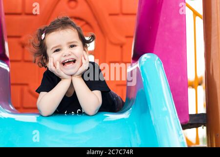 una ragazza liitle carina che si posa sullo scivolo blu in un parco giochi con le mani sotto il mento. Foto Stock