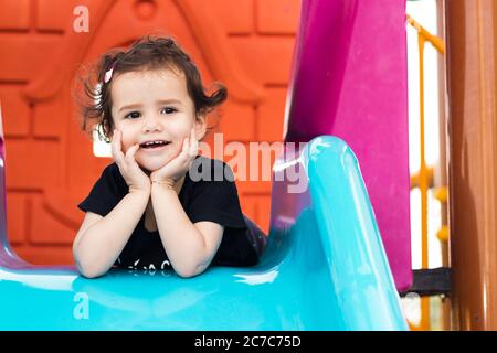 una ragazza liitle carina che si posa sullo scivolo blu in un parco giochi con le mani sotto il mento. Foto Stock