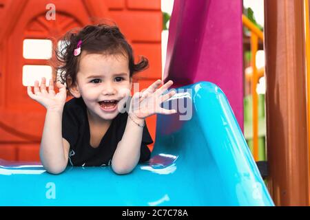 una ragazza liitle carina che si posa sullo scivolo blu in un parco giochi con le mani sotto il mento. Foto Stock