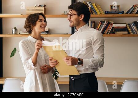 Giovani colleghi di affari attraenti che si levano in piedi con lettera della busta mentre felicemente si guardano l'un l'altro in ufficio moderno Foto Stock