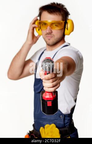 addetto alla riparazione caucasica in uniforme, occhiali di sicurezza e cuffie che tengono isolato il cacciavite senza fili su sfondo bianco. attenzione al cacciavite senza fili Foto Stock