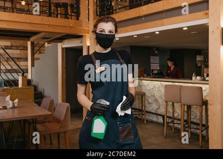 Una cameriera elegante che indossa un grembiule, una maschera medica nera e guanti medici monouso sta tenendo una bottiglia con disinfettante e un panno in un riposo Foto Stock