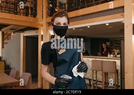 Un ritratto ravvicinato di una cameriera elegante che indossa un grembiule, una maschera medica nera e guanti medici monouso sta tenendo una bottiglia con disinfettante Foto Stock