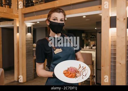 Una cameriera elegante che indossa una maschera nera e guanti monouso tiene un'insalata di menta e dolendwitz in un ristorante. Un barista carino sta servendo Foto Stock