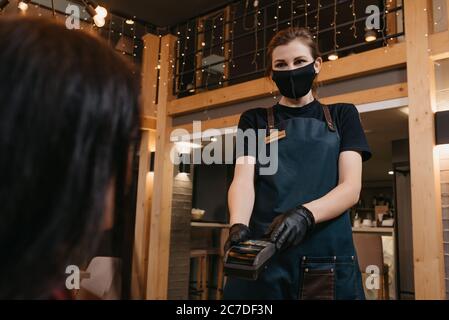 Una cameriera che indossa una maschera medica e guanti medici usa e getta sta consegnando un terminale di pagamento wireless a una cliente femminile in un ristorante. Il Foto Stock