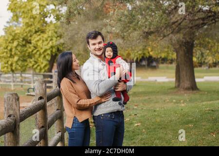 Felice madre cinese asiatica sorridente e padre caucasico con bambina in costume ladybug. Famiglia in autunno parco all'aperto. Halloween o grazie Foto Stock