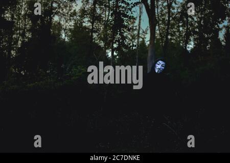 un maschio con cappuccio nero che indossa una maschera bianca in piedi sul sfondo di alberi in una foresta Foto Stock