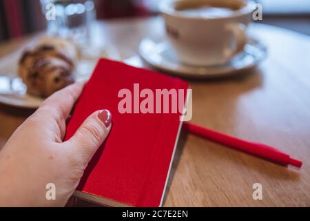 Una nota rossa e una penna rossa sono sul tavolo in un caffè, una tazza di caffè e un croissant sullo sfondo. Mano femmina che tiene un blocco note Foto Stock