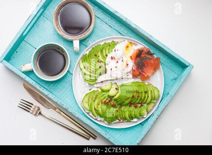 Bruschetta con salmone salato, avocado tritato, pepe e uovo in camicia su un piatto bianco su fondo turqoise Foto Stock