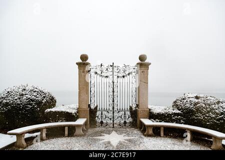 Un cancello chiuso in metallo nero con colonne di cemento circondate da piante verdi coperte di neve Foto Stock