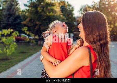 Felice madre che incontra figlia dopo le classi all'aperto scuola primaria e solletico bambino. Famiglia divertendosi ridendo Foto Stock